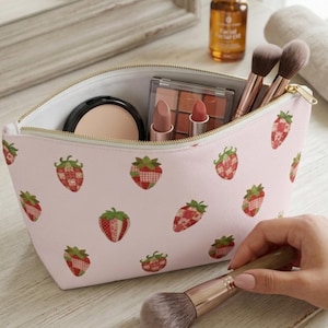 May include: A light pink cosmetic pouch featuring a repeating pattern of red and white patchwork-style strawberries with green leaves. The zippered bag is open, revealing makeup items including a compact, lipstick, and brushes inside.