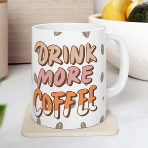 May include: A white ceramic mug with a handle featuring the text "DRINK MORE COFFEE" in stacked, stylised brown, pink, and orange letters. The mug is decorated with a repeating pattern of small brown coffee bean icons scattered across the surface.