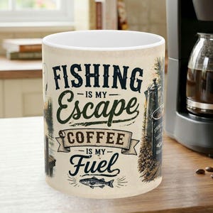 Peut inclure: Un mug en céramique blanche orné d'un motif graphique avec le texte Fishing is my Escape Coffee is my Fuel. Le design comprend des illustrations d'une scène de pêche, d'une cafetière et d'un poisson, dans des tons bleu foncé et marron.