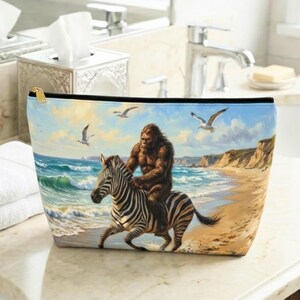 May include: A zippered pouch featuring a printed illustration of a brown, hairy creature riding a black and white zebra along a sandy beach. The scene includes blue ocean waves, a light blue sky with clouds, and several white seagulls flying overhead.