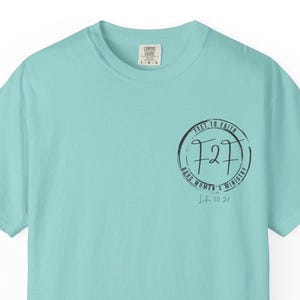 May include: A light teal Comfort Colors t-shirt with a circular graphic on the front. The graphic includes the text "FEET TO FAITH", "727", "BABC WOMEN'S MINISTRY", and "J.L. 10:27" in black ink. The shirt has a crew neck and short sleeves.