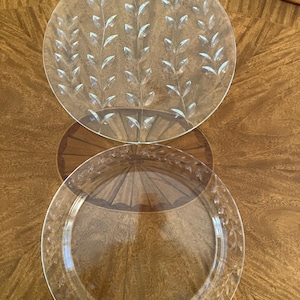 May include: Two clear glass plates. One plate features a leaf design etched into the glass. The other plate has a leaf design around the rim. Both plates are on a wooden table with a decorative pattern.