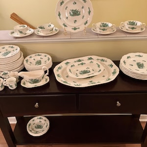 Op de afbeelding: Een verzameling crèmekleurig keramisch servies met een groen landschapsontwerp. De set bevat borden, kommen, kopjes, schotels en serveerschalen. De borden hebben geschulpte randen. Het servies is op een donkerbruine houten tafel geplaatst.