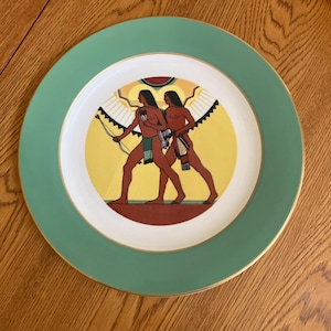 May include: A decorative plate with a green border and gold trim. The center features a yellow background with two figures in Native American attire, one holding a bow. The plate is a decorative item.
