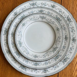 May include: A set of white porcelain plates with a delicate blue floral pattern and silver trim. The plates are arranged in a nested stack, showcasing the different sizes and designs. The plates are on a wooden surface.
