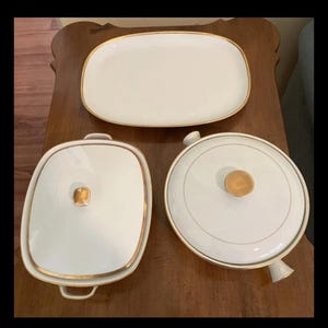 May include: A collection of white serving dishes with gold trim. The set includes a rectangular platter, a square lidded dish, and a round lidded casserole dish. The dishes are arranged on a wooden surface.