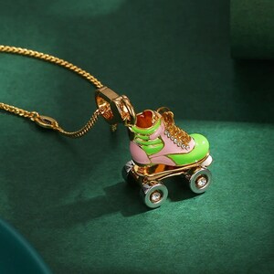 May include: A gold-colored necklace featuring a pendant shaped like a roller skate. The roller skate is painted in shades of pink and green, with gold accents and clear gem details on the wheels. The chain is also gold.