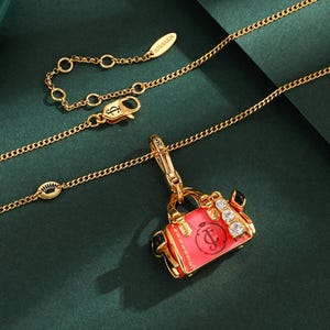 May include: Gold-toned necklace with a chain and a red handbag charm. The charm has a gold frame, black accents, and clear gemstones. The necklace has a lobster clasp and a small tag with text.