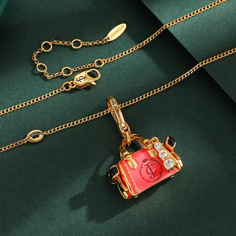 May include: Gold-toned necklace with a chain and a red handbag charm. The charm has a gold frame, black accents, and clear gemstones. The necklace has a lobster clasp and a small tag with text.