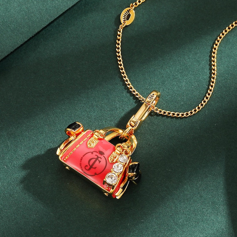 May include: Gold-toned necklace with a pendant shaped like a pink handbag. The handbag pendant features a black handle, sparkling rhinestones, and a cursive "J" logo. The necklace is on a dark green background.