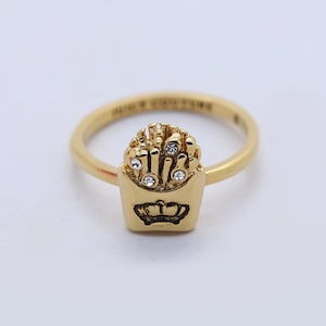 May include: A gold ring with a french fry design. The ring features a small, rectangular container of fries, decorated with a crown and sparkling details. The ring's band is thin and smooth, with a gold finish.