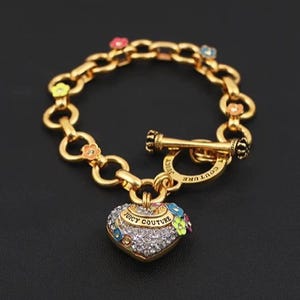 May include: Gold-coloured bracelet with a heart-shaped charm. The charm is encrusted with rhinestones and features the words "Juicy Couture". Small, colourful flowers and rhinestones adorn the bracelet's chain links.