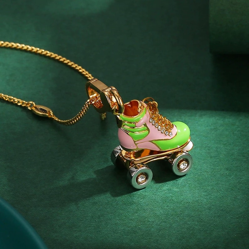 May include: A gold-colored necklace featuring a pendant shaped like a roller skate. The skate is painted in pink and green with gold accents and clear gem details on the wheels. The chain is a delicate gold color.