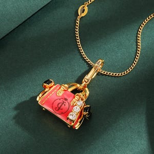 May include: Gold-toned necklace with a pendant shaped like a pink handbag. The handbag pendant features a black handle, sparkling rhinestones, and a cursive "J" logo. The necklace is on a dark green background.