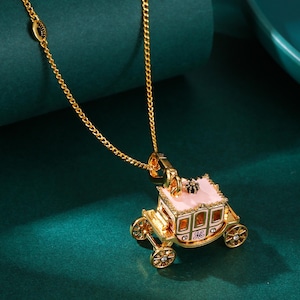 May include: A gold-colored necklace with a pendant shaped like a pink and gold carriage. The carriage has detailed windows and wheels, with a small black crown on top. The necklace chain is also gold and has a delicate design.