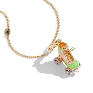 May include: A gold-colored necklace featuring a charm shaped like a roller skate. The skate is pink and green with silver wheels and a gold-colored lace. The necklace has a gold-colored chain and clasp.