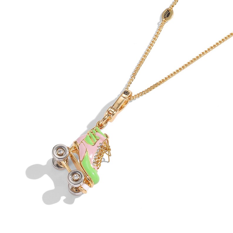 May include: A gold-colored necklace featuring a pendant shaped like a roller skate. The skate is pink and green with silver wheels and clear gem accents. The chain is delicate and has a lobster clasp.
