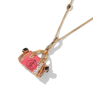 May include: Gold-toned necklace featuring a charm shaped like a pink handbag. The handbag charm has a black handle, clear rhinestones, and a black logo. The necklace chain is delicate and has a lobster clasp.