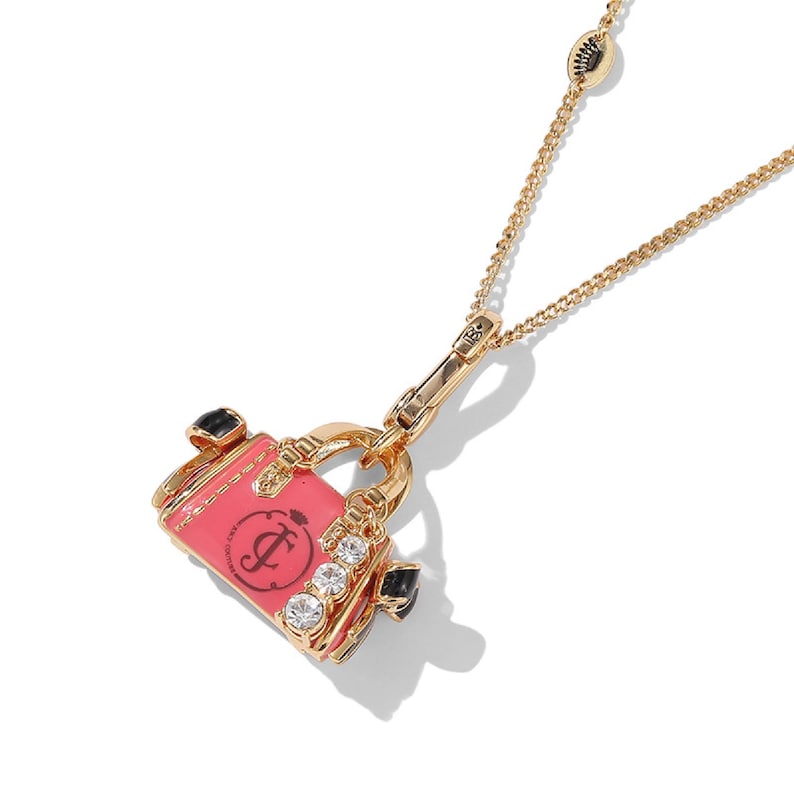 May include: Gold-toned necklace featuring a charm shaped like a pink handbag. The handbag charm has a black handle, clear rhinestones, and a black logo. The necklace chain is delicate and has a lobster clasp.