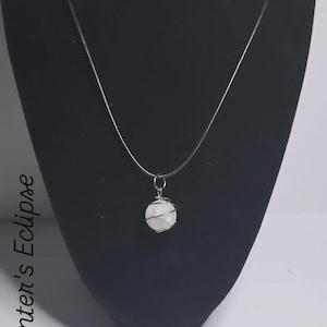 May include: A silver necklace featuring a round, white crystal pendant wrapped in silver wire. The pendant hangs from a delicate silver chain. The words "Winter's Eclipse" are printed vertically on the left side of the image.