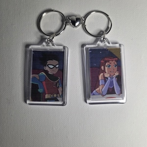 May include: Two keychains featuring cartoon characters. One keychain shows a character in a red and blue outfit with a black mask. The other keychain shows a character with purple and blue clothing. The keychains are connected by a heart-shaped clasp.