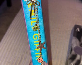 May include: A gold chain bookmark with various charms, including a bow, a green gem, a starfish, and a smiley face, hangs from a book with a turquoise cover. The book's title is partially visible, with the name "GRIFFIN" in yellow letters.