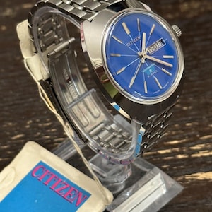 May include: A vintage Citizen automatic wristwatch with a bright blue dial. The watch has a silver-tone stainless steel strap and case. The dial shows the day and date, with the brand name and "21 Jewels" visible. The watch is displayed on a clear stand.