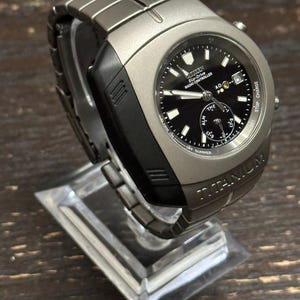 May include: A titanium Citizen Eco-Drive radio-controlled watch with a black face. The watch has a silver-tone case and band, and is displayed on a clear stand. The watch face includes multiple sub-dials and the word "TITANIUM".