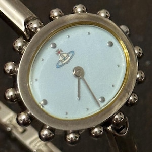 May include: A silver-toned wristwatch featuring a pale blue circular dial with a small orb logo at the twelve o'clock position. The watch case is surrounded by a decorative border of small silver spheres and is attached to a matching metal band.