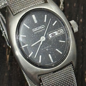 Op de afbeelding: Een vintage Seiko automatisch horloge met een donkergrijze wijzerplaat, zilveren wijzers en een datumweergave. Het horloge heeft een zilveren mesh band en een zilveren kast. De wijzerplaat toont de tekst "SEIKO AUTOMATIC".