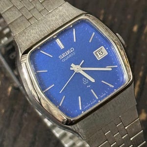 May include: A silver-tone Seiko Quartz wristwatch featuring a square case with a vibrant blue dial. The watch includes silver hour markers, silver hands, and a date window at the three o'clock position. It has a matching silver-tone metal link bracelet.