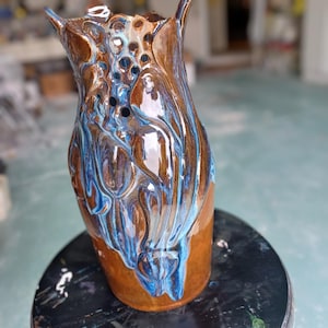 Può includere: Un vaso in ceramica fatto a mano con una forma organica unica. Il vaso presenta una ricca base marrone, che sfuma in una superficie lucida con smalto blu e bianco fluente. La parte superiore ha un design strutturato e aperto. Un pezzo decorativo per la casa.