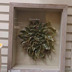 Pode incluir: Escultura floral em cerâmica emoldurada. A obra apresenta uma flor verde brilhante e detalhada com múltiplas camadas de pétalas. A flor está envolta em uma moldura de madeira marrom clara com uma frente de vidro, criando um efeito de caixa de sombra. O fundo é bege neutro.