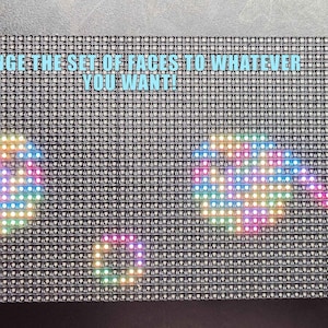 May include: A rectangular LED display with a grid of small, dark gray squares. The display shows three colorful, pixelated shapes resembling faces. Text at the top reads "CHANGE THE SET OF FACES TO WHATEVER YOU WANT!" A USB cable is plugged into the side.
