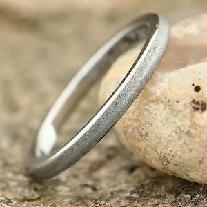 May include: A silver ring with a textured surface. The ring is set against a neutral background, highlighting its simple design. The ring's band appears to be approximately 2mm wide.