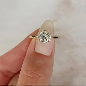 Könnte beinhalten: Ein goldener Verlobungsring mit einem großen, runden, klaren Edelstein. Der Ring hat ein schlichtes Band und eine sechs-Zacken-Fassung. Der Edelstein ist der Mittelpunkt und reflektiert das Licht.