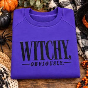 May include: A vibrant purple crewneck sweatshirt featuring the black embroidered text WITCHY, OBVIOUSLY. The garment has long sleeves, a ribbed collar, and a soft fleece-like texture, presented as a solid-colored pullover top.