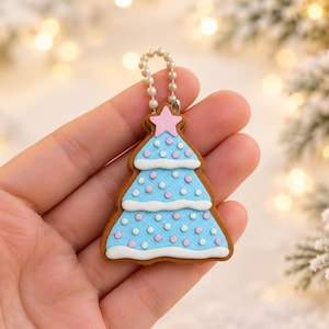 Puede incluir: Un adorno con forma de árbol de Navidad con borde marrón estilo pan de jengibre, capas azul claro texturizadas, ribete blanco festoneado y pequeños puntos rosas y blancos. Una pequeña estrella rosa corona la parte superior con una cadena.
