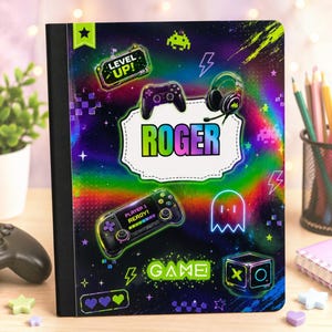 May include: A rectangular notebook with a black spine and a vibrant, holographic-style cover featuring gaming graphics. The cover displays the name ROGER in large letters, alongside illustrations of game controllers, a headset, and the text LEVEL UP.