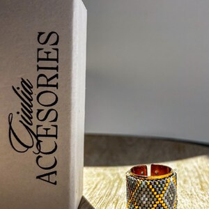 May include: A beaded ring with a copper-colored band and a geometric pattern in gray, yellow, and teal. The ring is next to a box with the text "Giulia ACCESORIES". The ring is on a wooden surface.