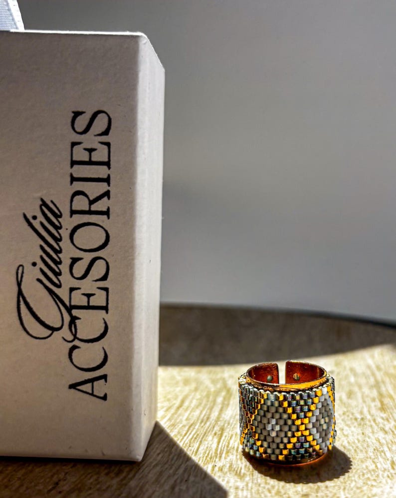 May include: A beaded ring with a copper-colored band and a geometric pattern in gray, yellow, and teal. The ring is next to a box with the text "Giulia ACCESORIES". The ring is on a wooden surface.