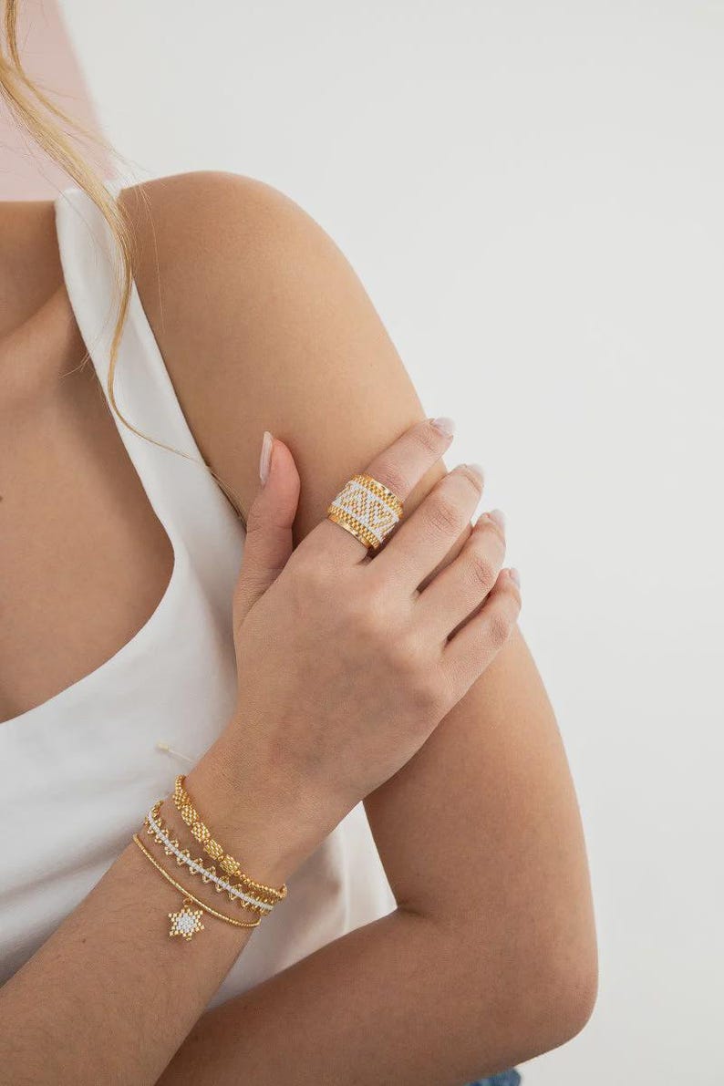 May include: A gold-toned ring with a woven design and a matching gold bracelet set. The ring features a band of white stones. The bracelets include a star charm. The jewelry is displayed on a person wearing a white top.