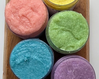 May include: An overhead shot of five small, clear jars filled with colorful, textured body scrub. The scrubs are pink, yellow, green, blue, and purple. The jars are arranged on a wooden cutting board.