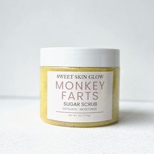 Op de afbeelding: Een pot gele suikerscrub met een witte deksel. Het etiket luidt "SWEET SKIN GLOW MONKEY FARTS SUGAR SCRUB EXFOLIATE, MOISTURIZE" en "Netto gewicht 6oz (170g)". De pot staat tegen een witte achtergrond.