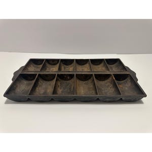 Puede incluir: Una sartén rectangular de hierro fundido con 16 compartimentos individuales. La sartén es de color marrón oscuro con una superficie texturizada y bordes redondeados. Los compartimentos están dispuestos en dos filas de ocho.