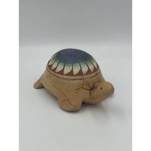 May include: A ceramic turtle figurine with a tan body and a shell painted with blue, white, and brown geometric patterns. The turtle has a rounded shape and is likely a decorative item.