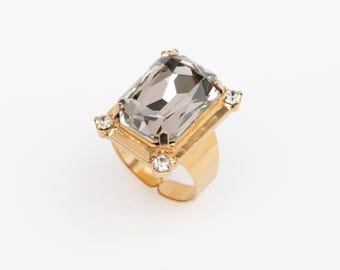 Könnte beinhalten: Ein goldfarbener Ring mit einem großen, rechteckigen, grauen Edelstein. Der Edelstein ist in einem goldenen Rahmen mit kleinen, klaren Steinen an jeder Ecke gefasst. Der Ring hat einen verstellbaren Ring.