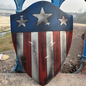 Puede incluir: Un escudo con una sección superior azul con tres estrellas plateadas. La sección inferior tiene rayas verticales rojas y blancas. El escudo tiene un aspecto desgastado y envejecido, con sombras oscuras en los bordes.