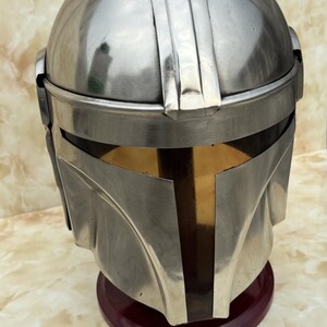 Puede incluir: Un casco plateado met&aacute;lico con una visera distintiva y una parte superior redondeada. El casco se exhibe sobre un soporte rojo oscuro, mostrando su superficie pulida y su intrincado dise&ntilde;o. El dise&ntilde;o del casco recuerda una est&eacute;tica espacial.