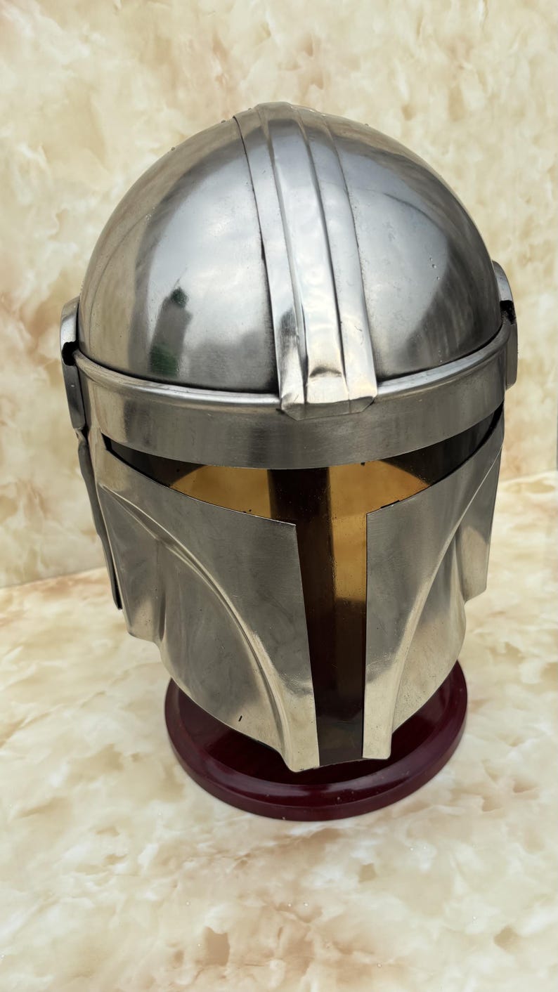 Puede incluir: Un casco plateado met&aacute;lico con una visera distintiva y una parte superior redondeada. El casco se exhibe sobre un soporte rojo oscuro, mostrando su superficie pulida y su intrincado dise&ntilde;o. El dise&ntilde;o del casco recuerda una est&eacute;tica espacial.